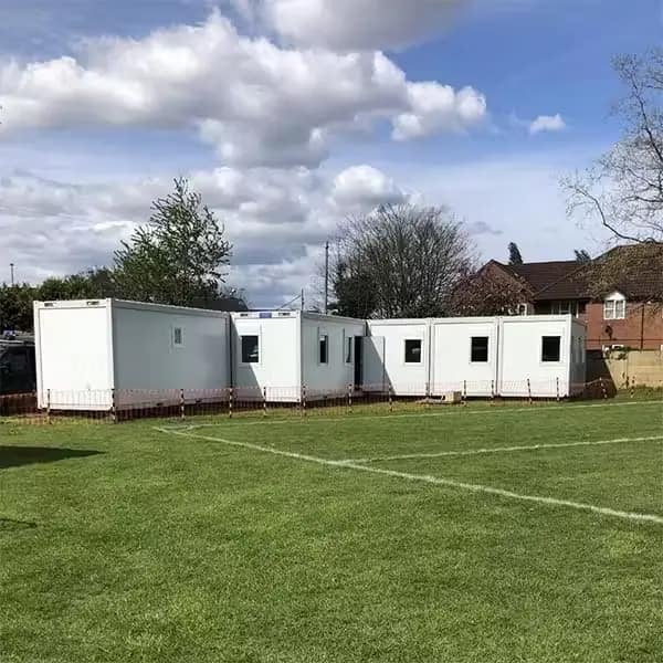 White modular classrooms arranged in a row on a green field with a cloudy sky overhead.