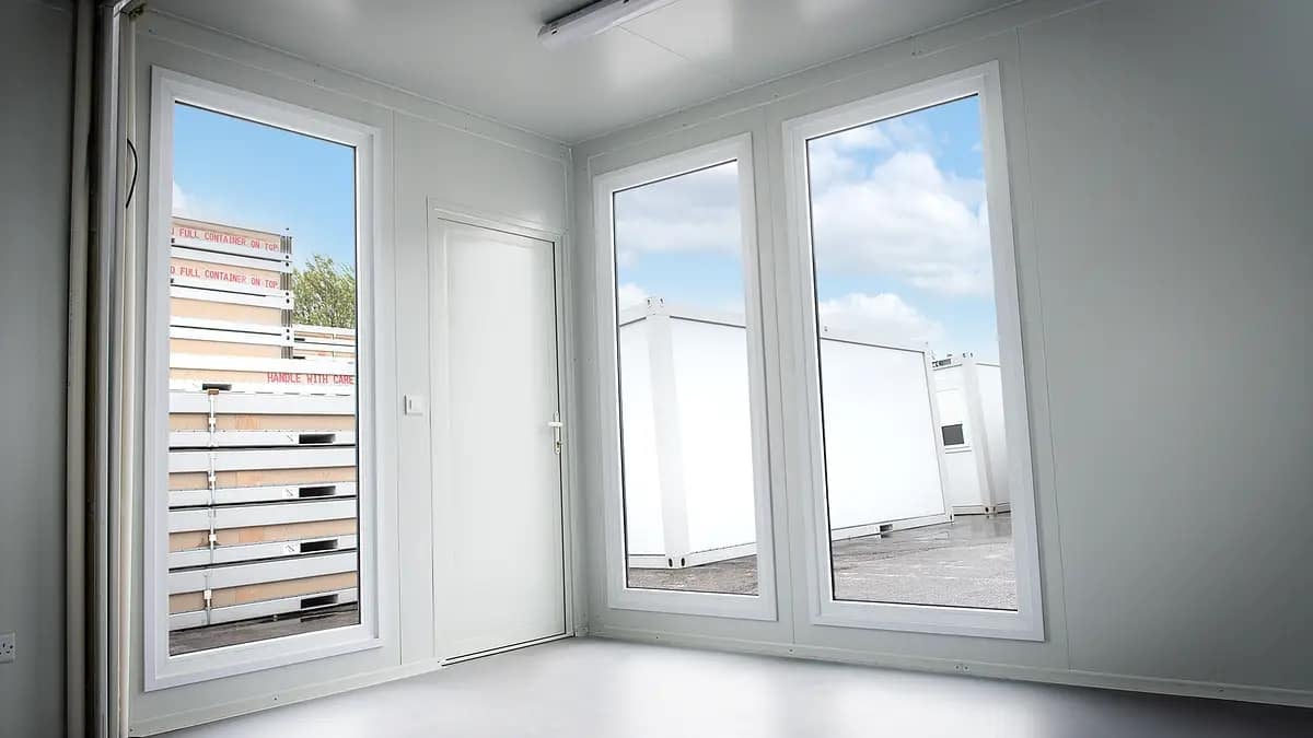 Bright, empty interior room with large windows overlooking stacked container units outside.