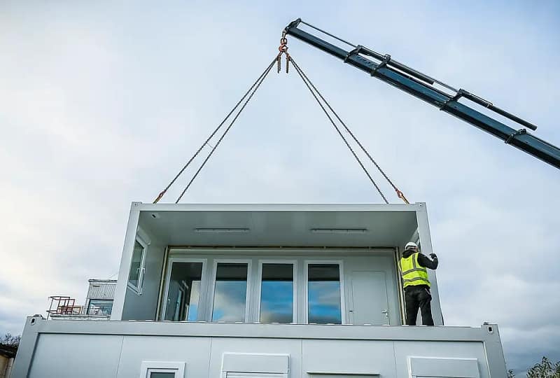 A crane lifting a prefabricated modular section of a building with a worker in high-visibility clothing guiding it into place against a cloudy sky backdrop.