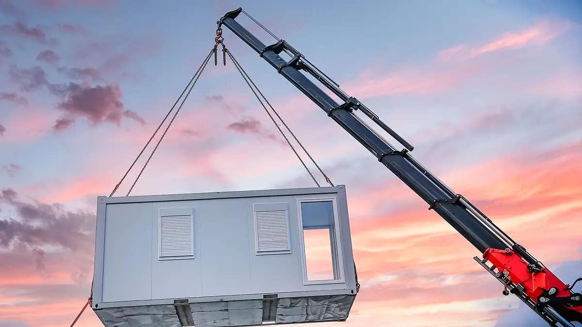 "Alt: A crane lifting a modular building unit against a sunset sky with pink and blue hues