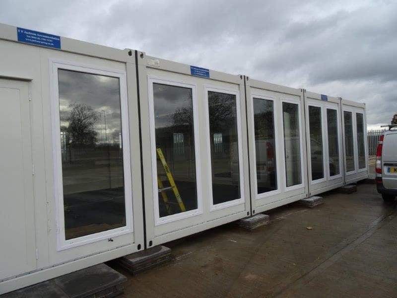 A series of large windows on a modular building, possibly a portable office, with reflections of a cloudy sky